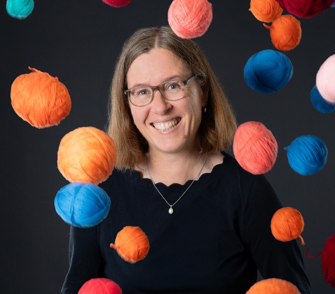Die Autorin Leonie Kramer lacht in die Kamera, um sie herum fliegen bunte Wollknäuel durch die Luft