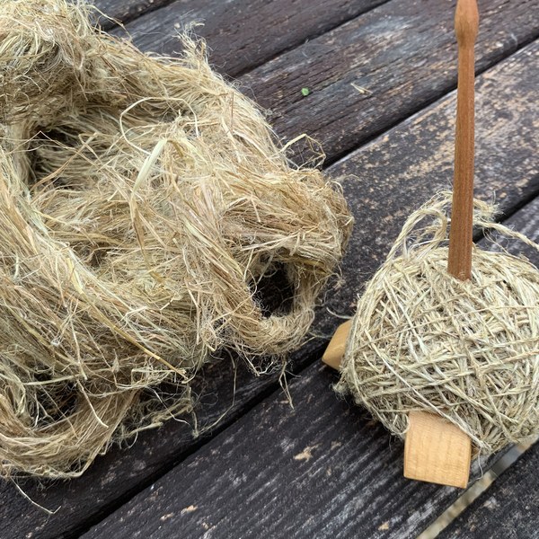 Eine Handspindel aus Holz mit aufgewickeltem, hellbraunem
                                   Brennnesselfaden, daneben ein Nest aus aufbereiteten Fasern