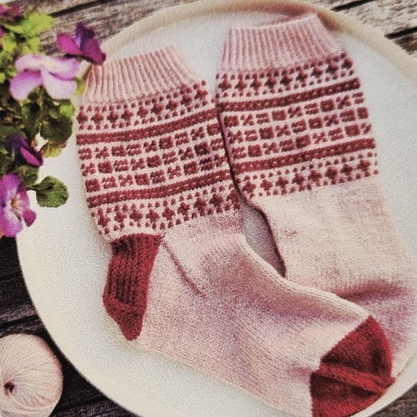 Ein Paar handgestrickte Socken in Altrosa und Dunkelrot auf einem hellen
                                   Teller: der Schaft trägt ein zweifarbiges Colourwork-Muster, Ferse und
                                   Spitze sind dunkelrot abgesetzt