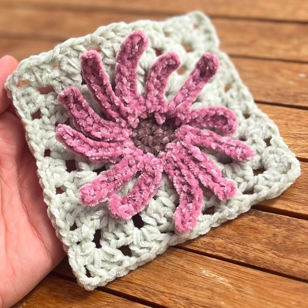 Ein gehäkeltes Granny Square aus flauschigem Chenillegarn: eine rosa Blüte
                                   mit vielen Blütenblättern auf hellgrauem Grund, in einer Hand gehalten